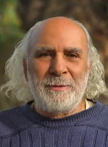 An elderly man with long white hair and a beard, wearing a dark sweater, outdoors with blurred greenery in the background.
