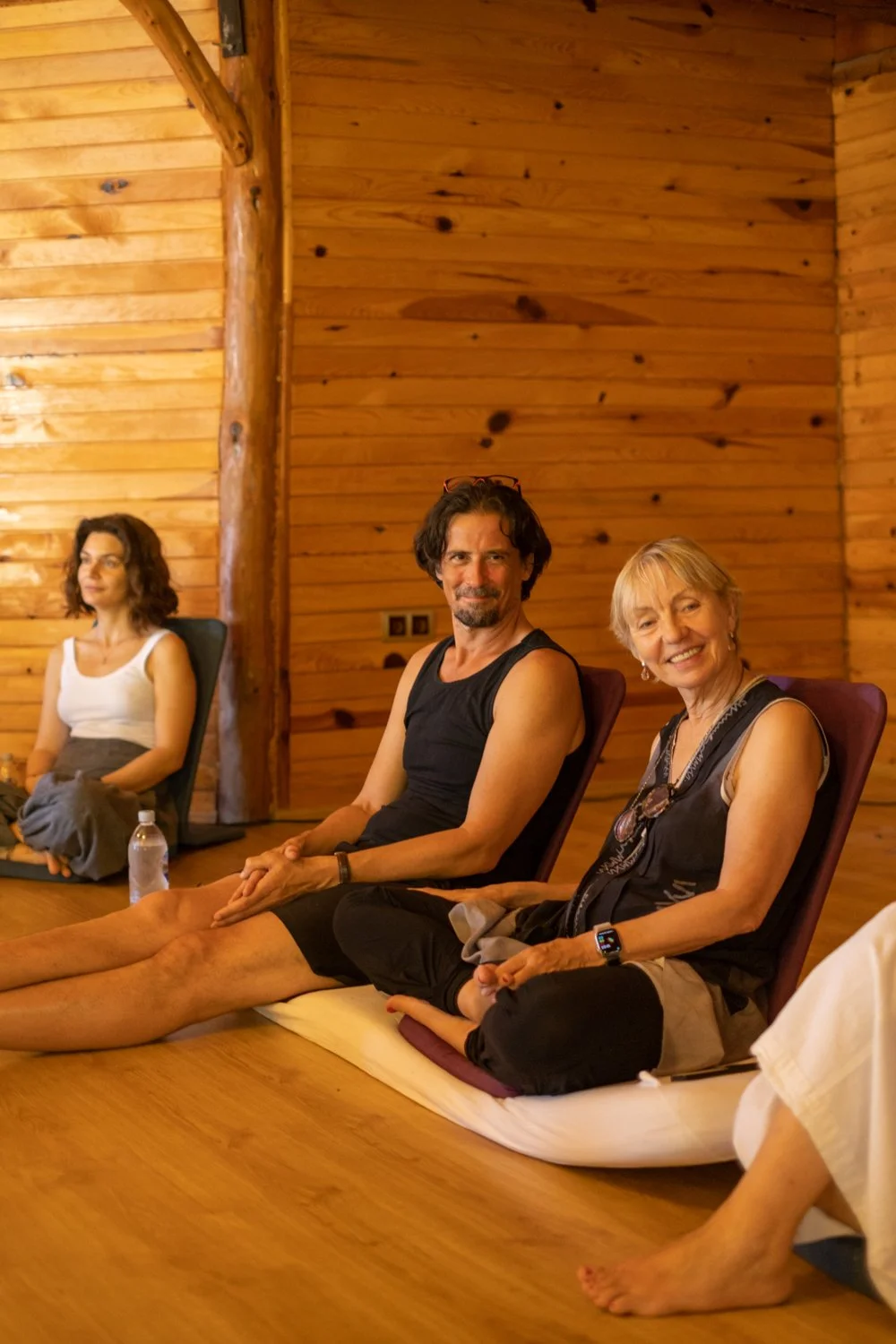 Three people sit on the floor in a wooden room, with the woman on the right smiling and wearing a watch, the man in the middle looking at the camera, and the woman on the left sitting back with her eyes closed.