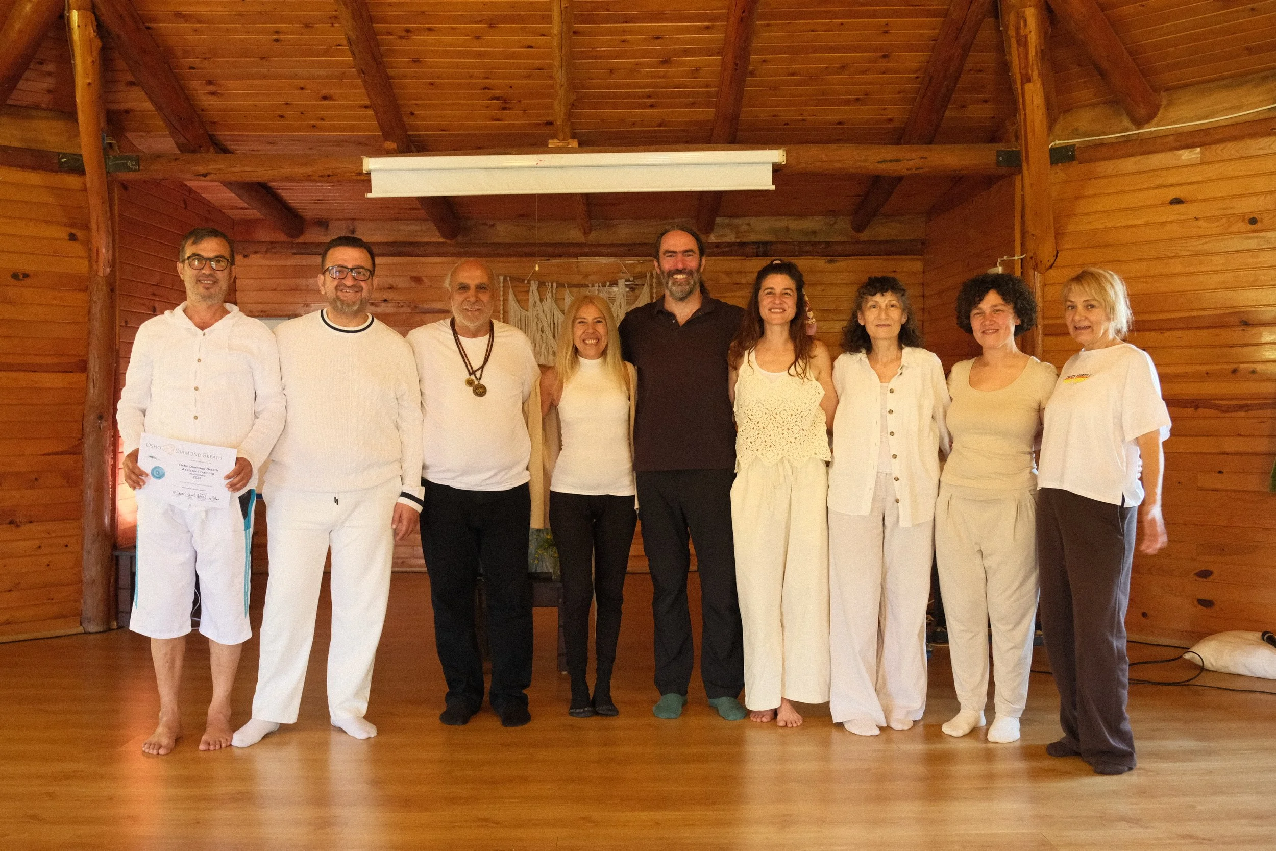 Group of nine people standing together indoors in front of wooden walls, smiling at the camera. Some are dressed in white clothing. Osho celebration, evening meeting, diamond breath school.