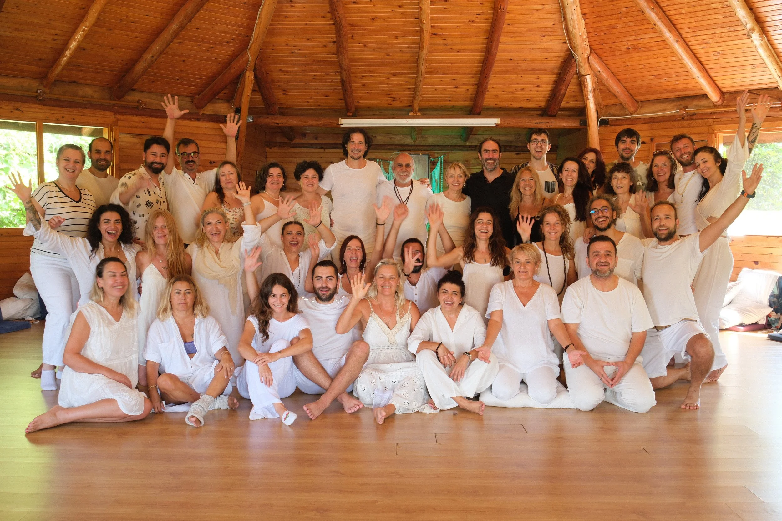 A large group of smiling people gathering together in a wooden room, some sitting and some standing, with many raising their hands in the air and waving. Osho celebration, evening meeting, diamond breath school.