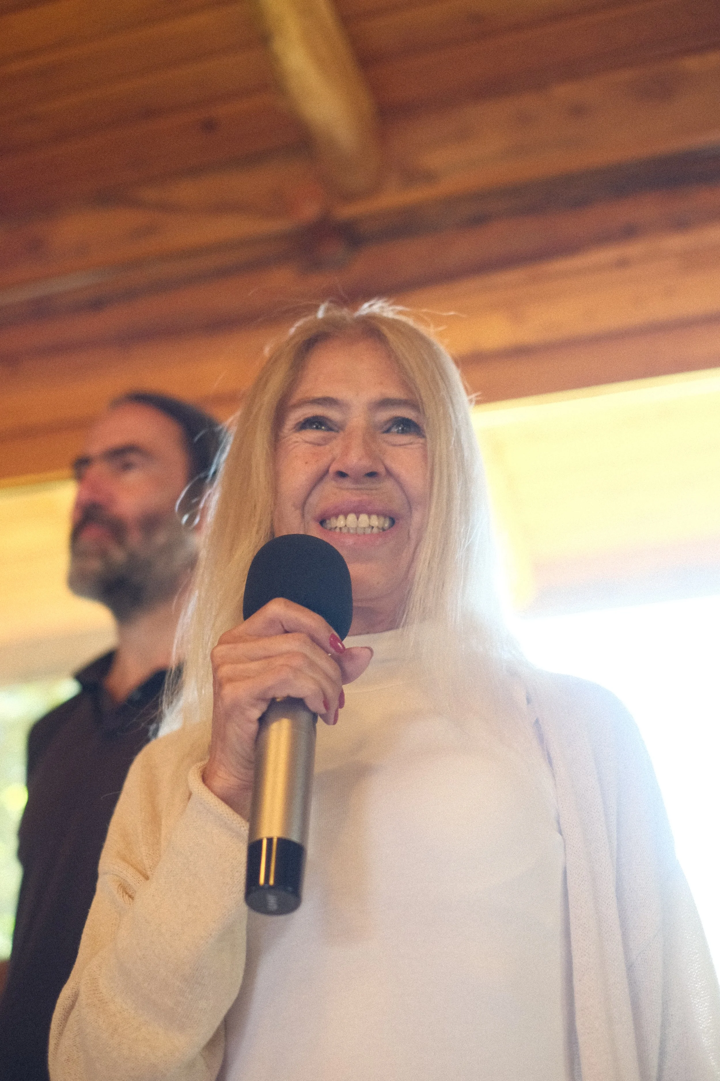 A woman with long blonde hair smiling and holding a microphone, with a man in the background, inside a room with wooden ceiling beams and warm lighting.