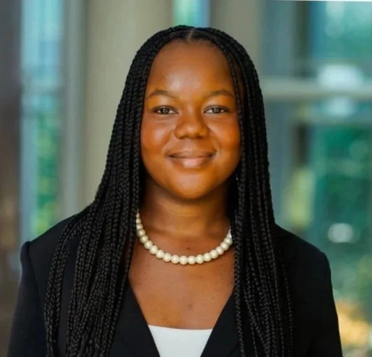 Portrait of a smiling woman with braided hair, wearing a pearl necklace and a black blazer, standing indoors with a blurred background.