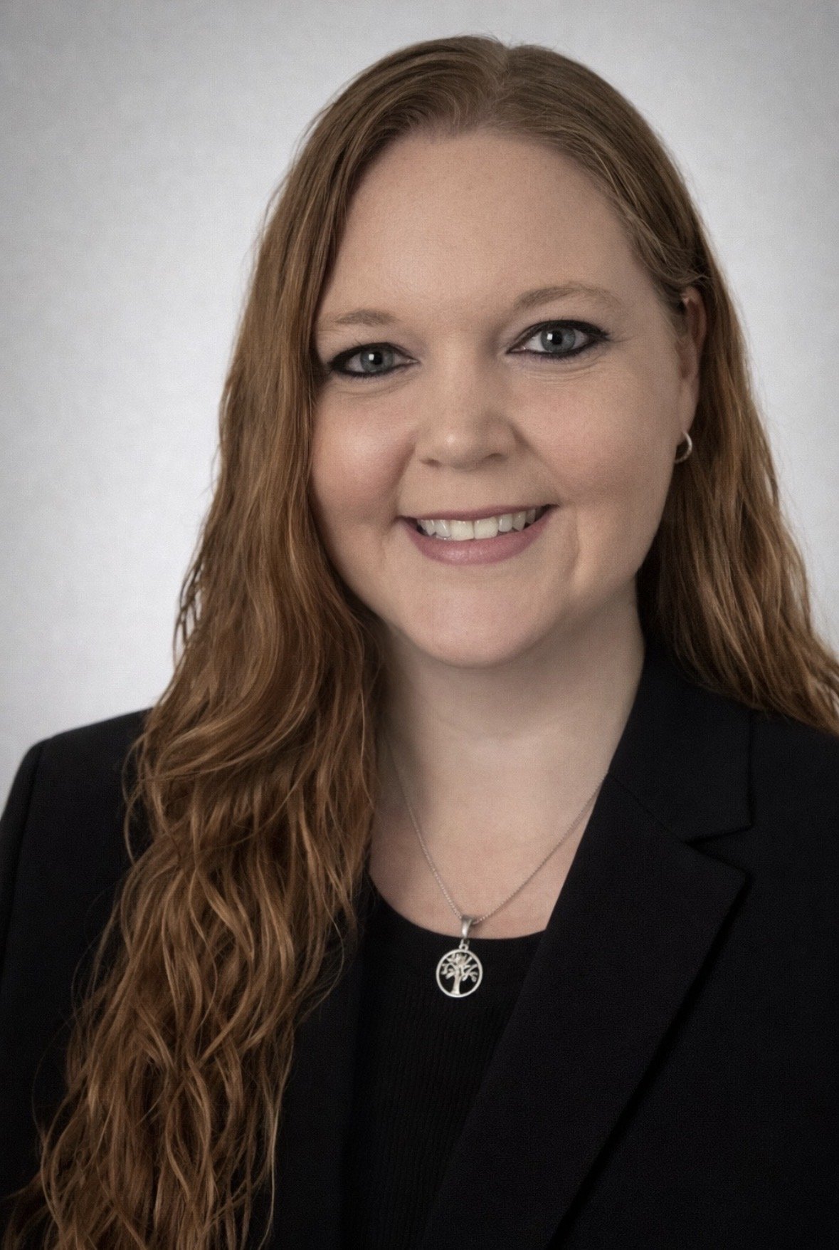 A woman with long, curly red hair, blue eyes, and wearing a black blazer and a pendant necklace smiling at the camera.