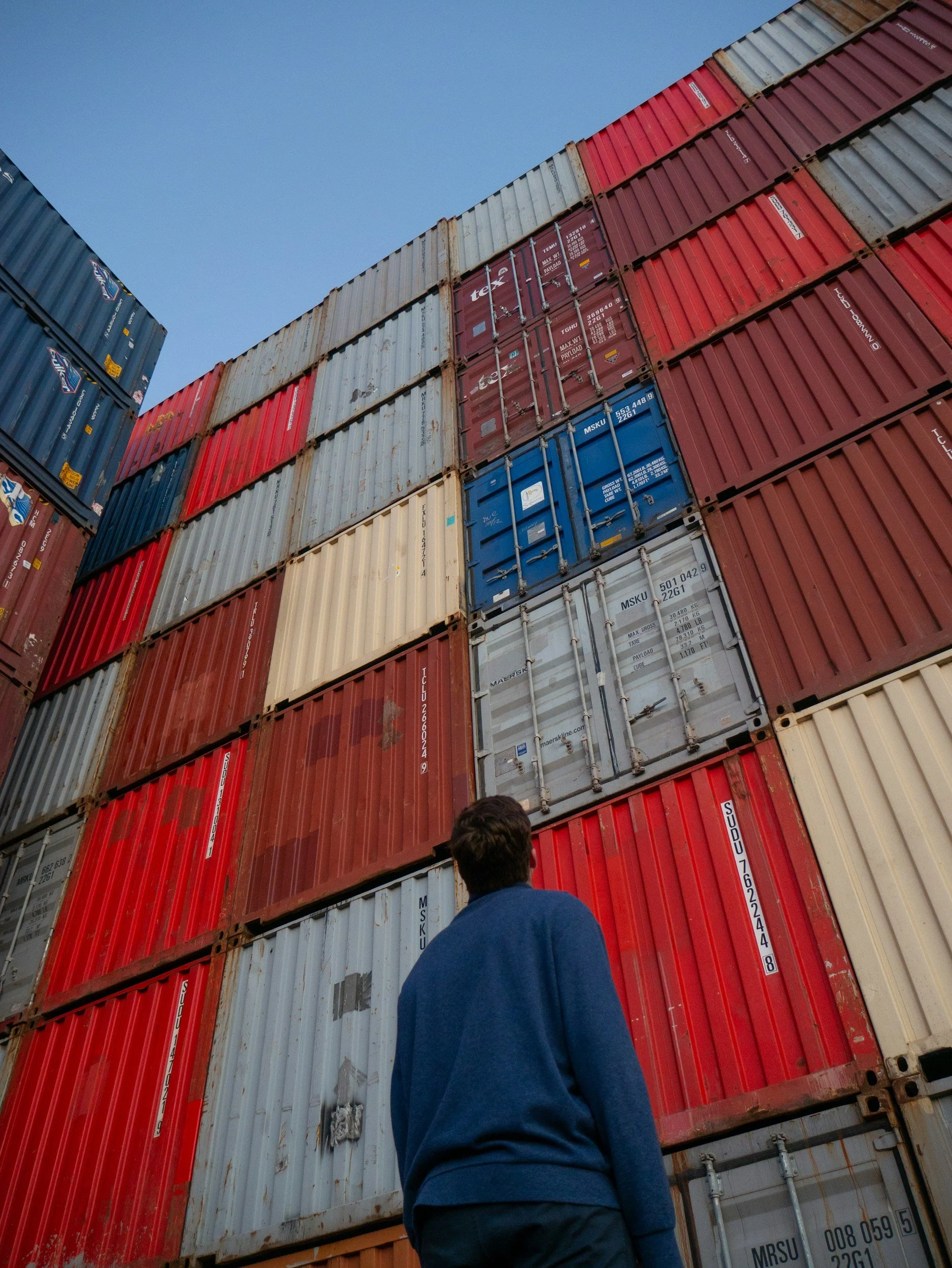 A person wearing a blue shirt standing in front of a large stack of colorful shipping containers, looking up at them under a clear blue sky.