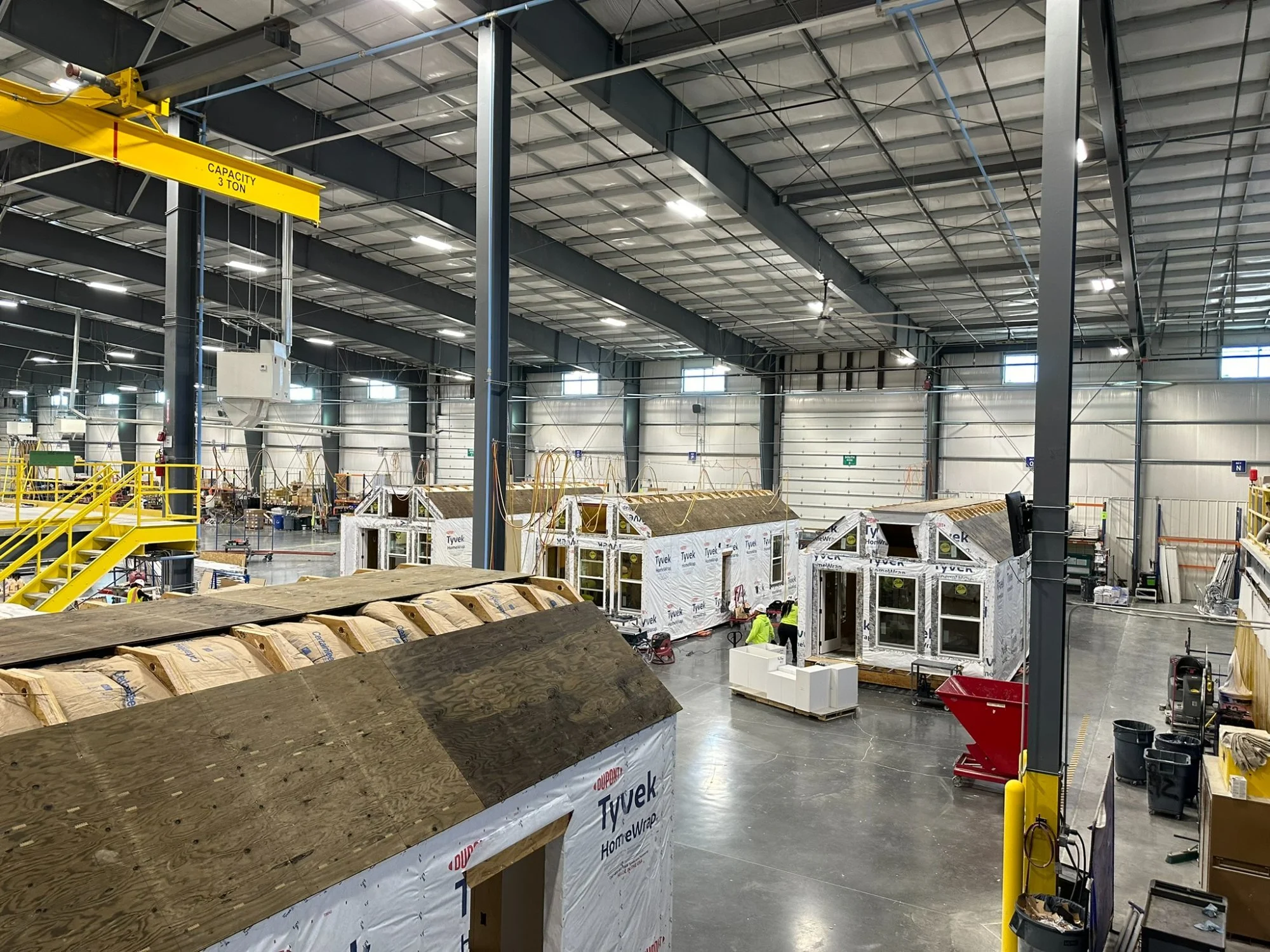 Interior view of Fading West Factory  steel beams, insulation, and electrical wiring. There is a yellow overhead crane with a capacity of 3 tons. Several small structures are being built, and workers are present.