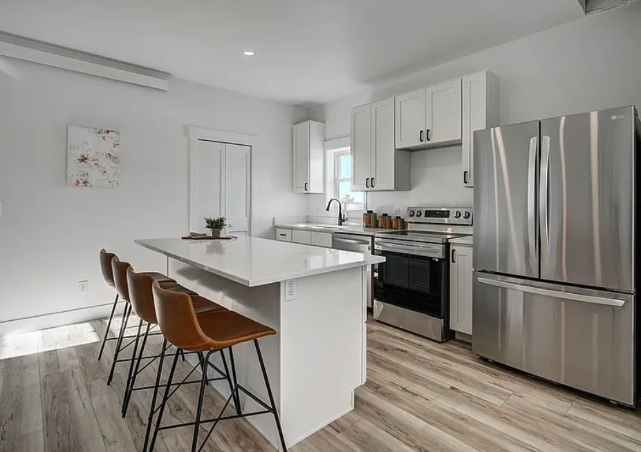 Interior photo of a Fading West Modular single-family home featuring a Modern kitchen with white cabinets, stainless steel refrigerator, black oven, island with brown chairs, and a small window with natural light.
