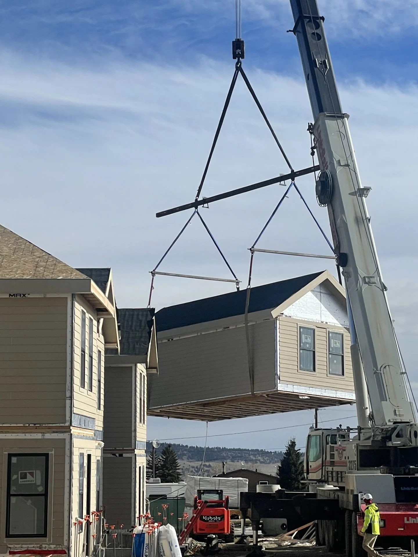 A construction crane lifting a modular house section onto a Fading West building site with residential houses and trees in the background.
