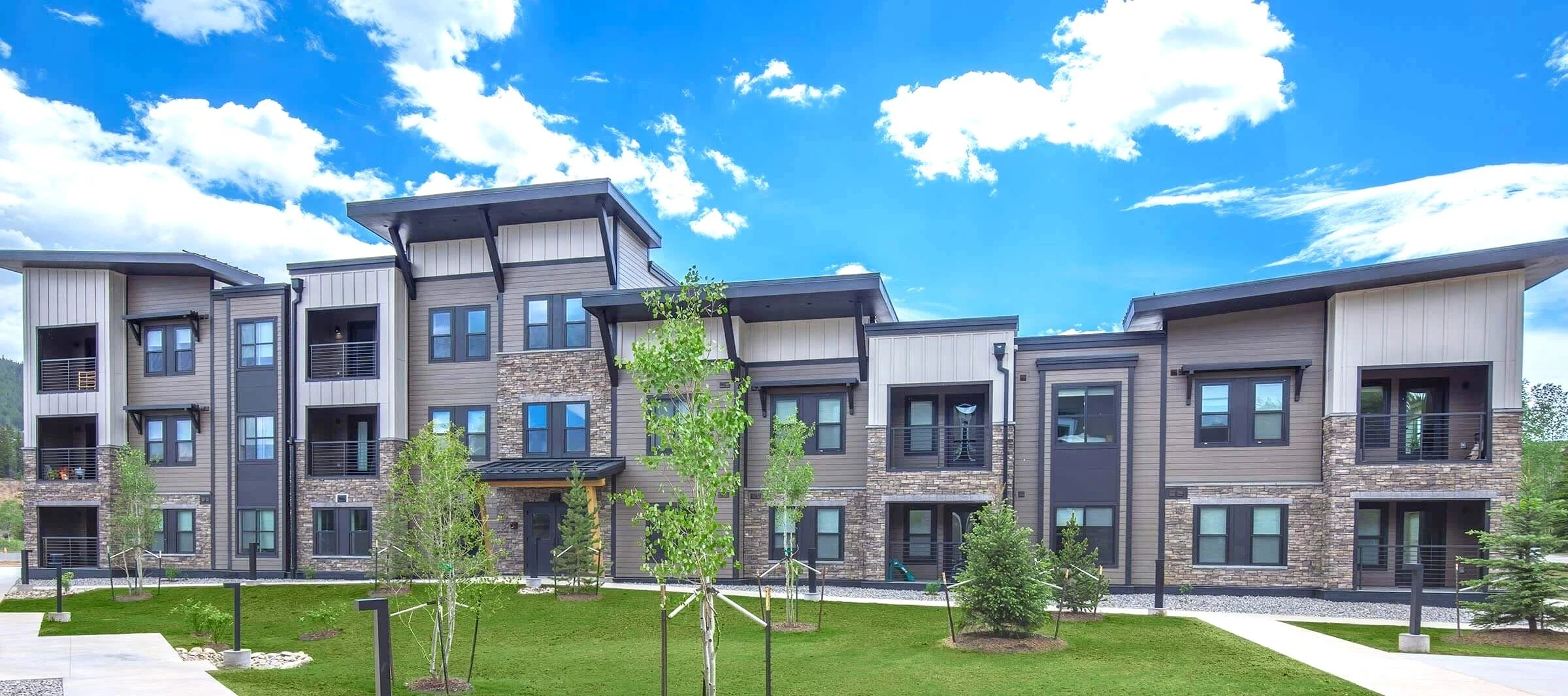 Modern multi-story modular apartment building with stone accents, metal balconies, and large windows, surrounded by young trees and a landscaped green lawn under a partly cloudy blue sky.