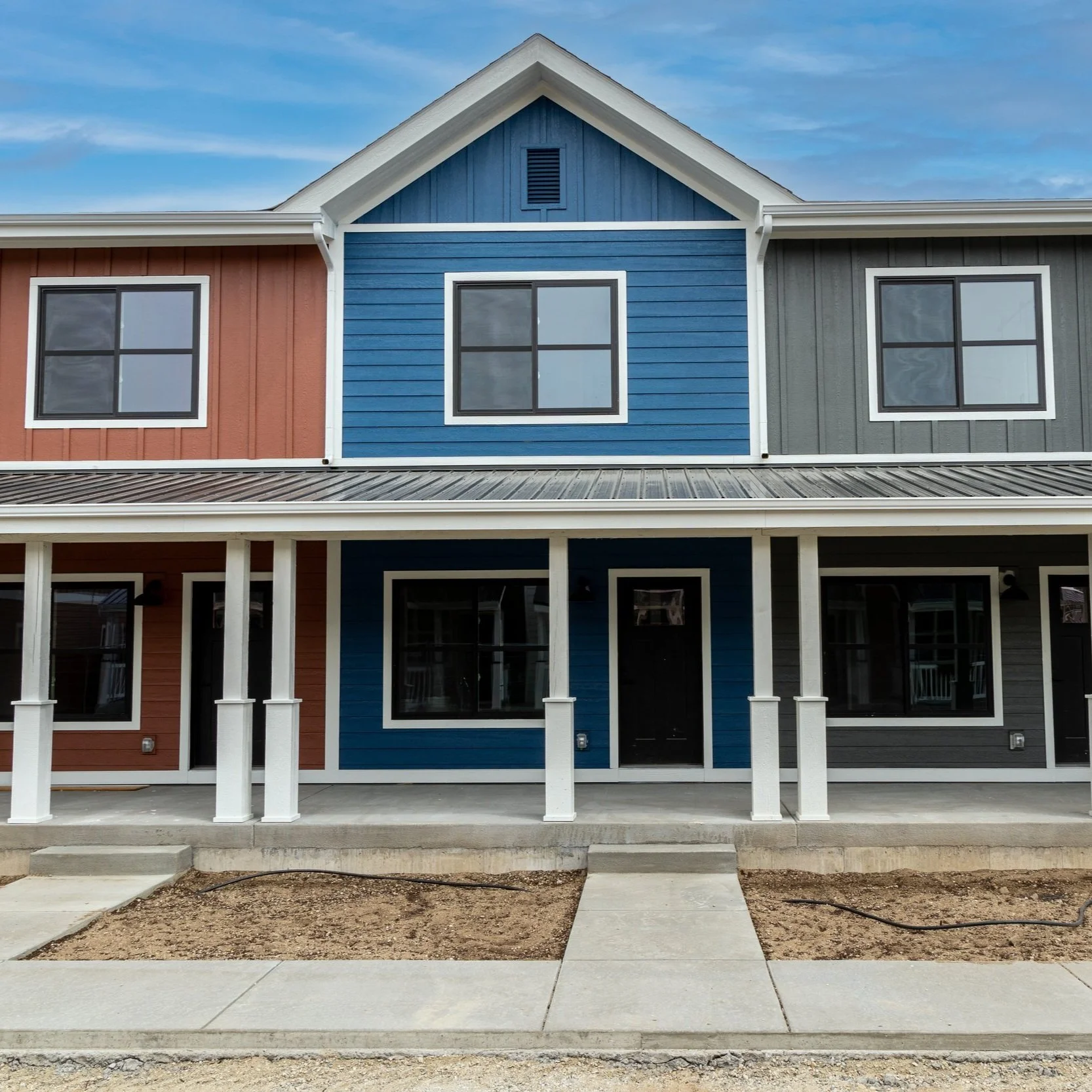 Fading West modular apartment building with three units featuring brown, blue, and gray exterior, covered porch with white columns, and concrete walkway.
