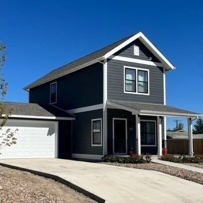 A modern two-story modular house by Fading West painted in dark gray with white trim, featuring a front porch, a garage, and a bright blue sky.