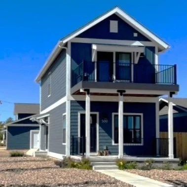 Two-story blue modular house by Fading West with white trim, front porch, and balcony under a blue sky.