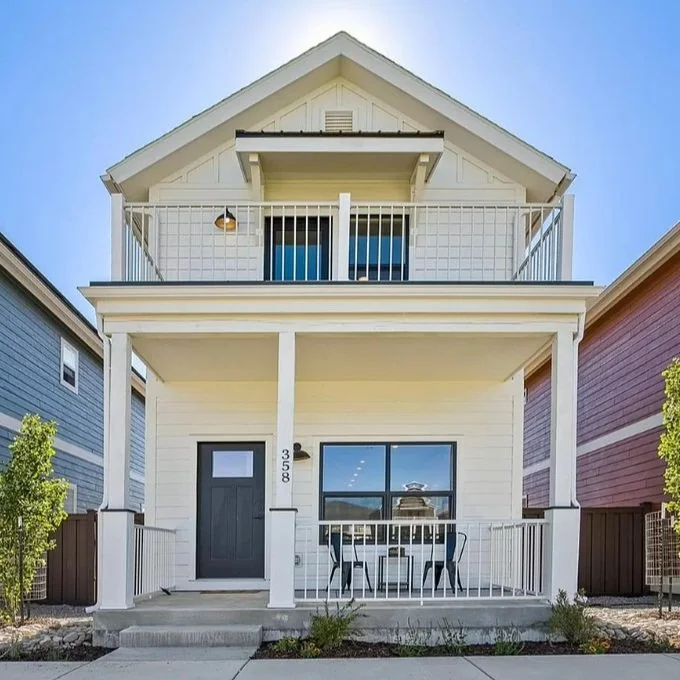 Front view of a two-story modular home by Fading West white house with a balcony and black front door, located between two colorful neighboring houses.