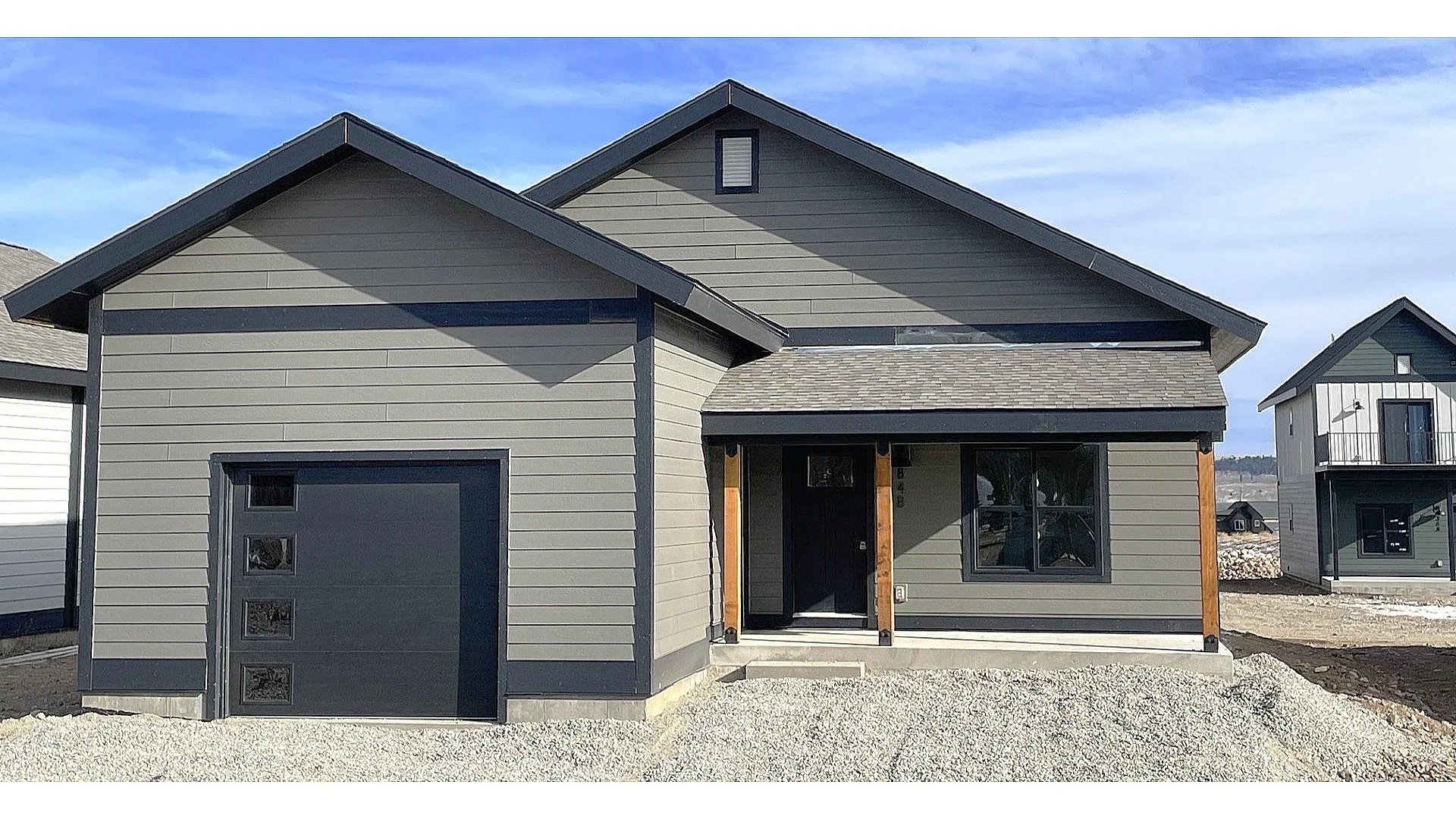 Fading West two-story single family modular house mid-installation — gray siding, black garage door, and covered porch with neighboring modular homes visible in background.