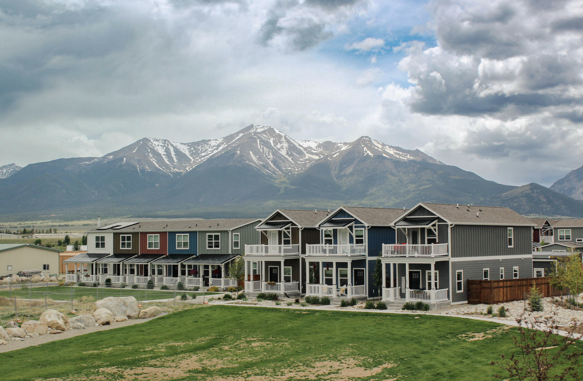 Colorful row of modern modular houses with large front windows and porches, set in a neighborhood with mountains and cloudy sky in the background.