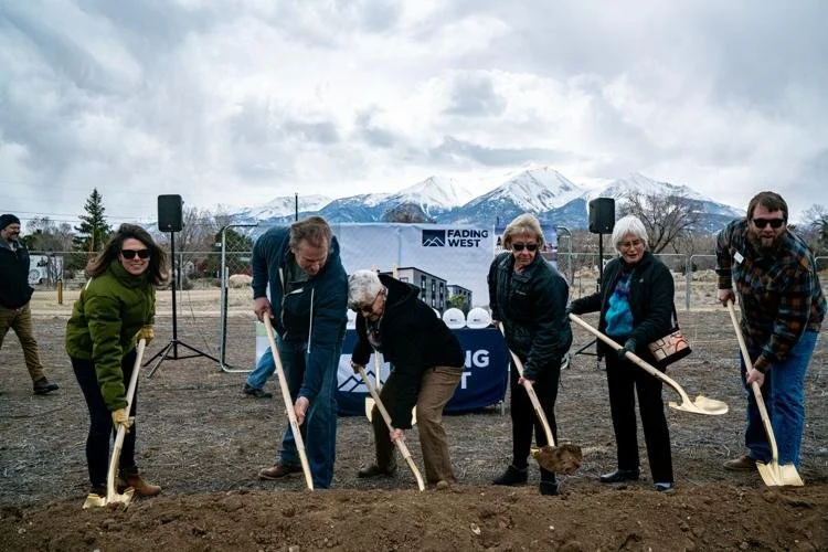 Group of six people at a groundbreaking workforce housing modular housing event, holding shovels in front of dirt with mountains in the background and a banner reading 'Fading West' in the distance.