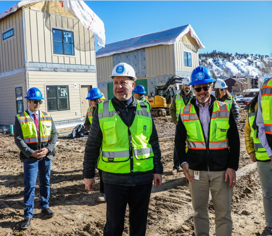 Modular Housing Fading West Construction site with workers and officials wearing safety vests and helmets, new houses being built in the background, snowy hills, and clear blue sky.