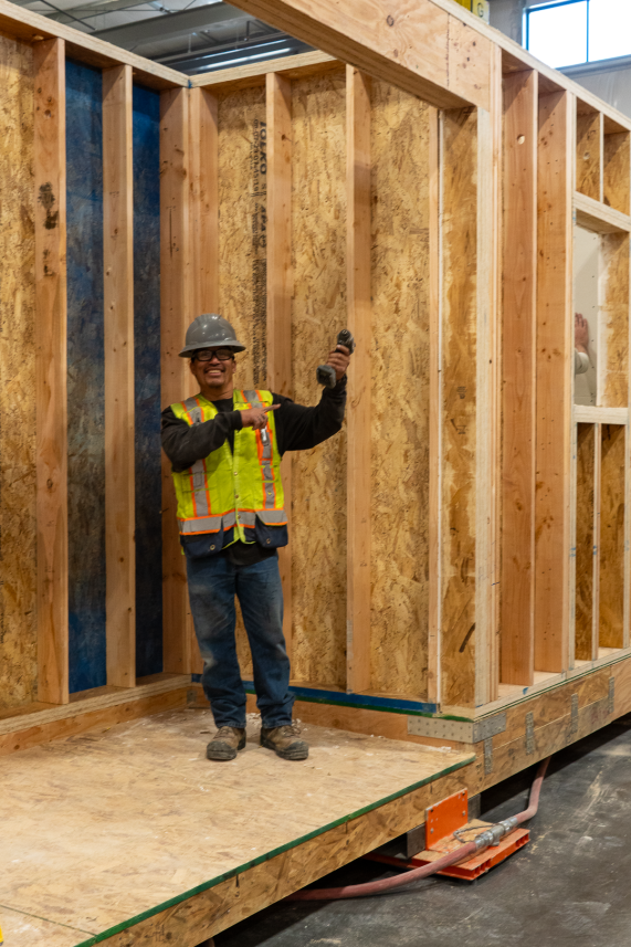 A Fading West construction worker wearing a yellow safety vest, hard hat, and glasses, smiling and holding a power drill inside a wooden framed structure under construction.