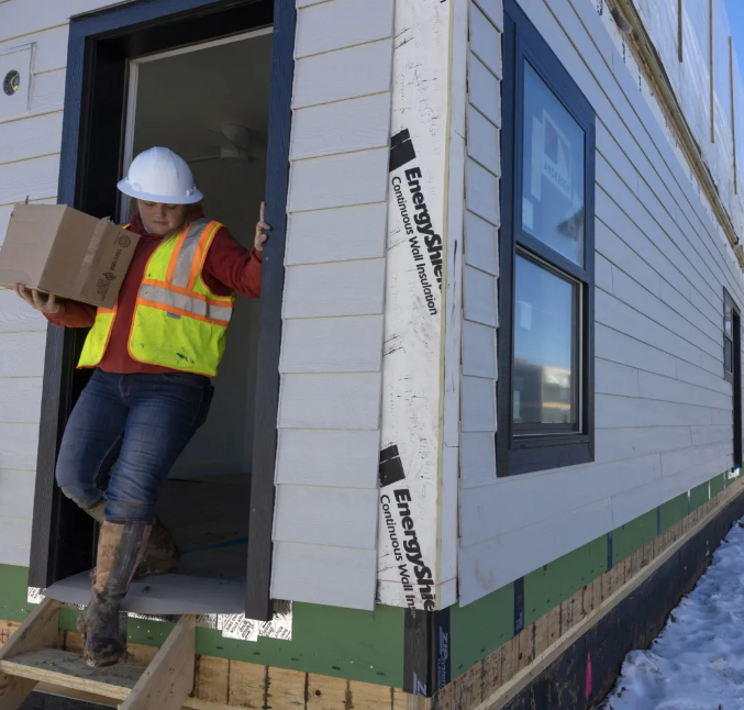 A Fading West Modular construction worker wearing a safety vest and hard hat carrying a box, standing in the doorway of a new, unfinished house under construction.