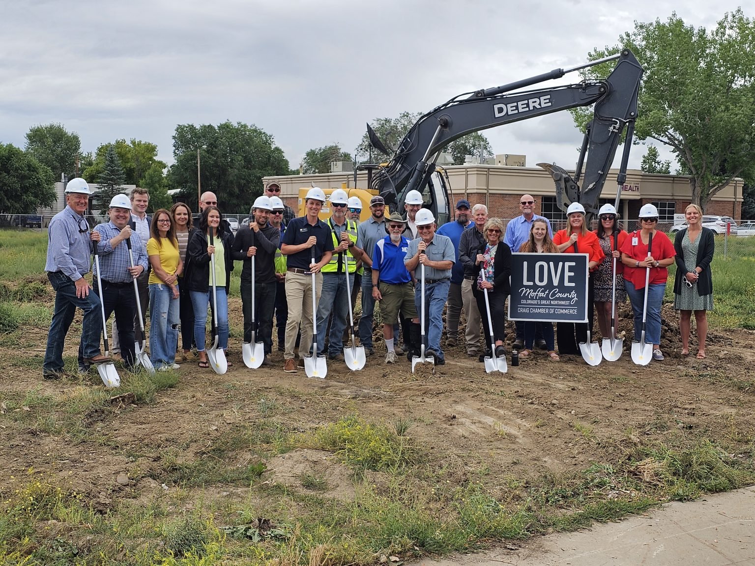 Fading West workforce housing groundbreaking ceremony in Moffat County, Colorado — large group in safety helmets with shovels, excavator, and Craig Chamber of Commerce sign in background