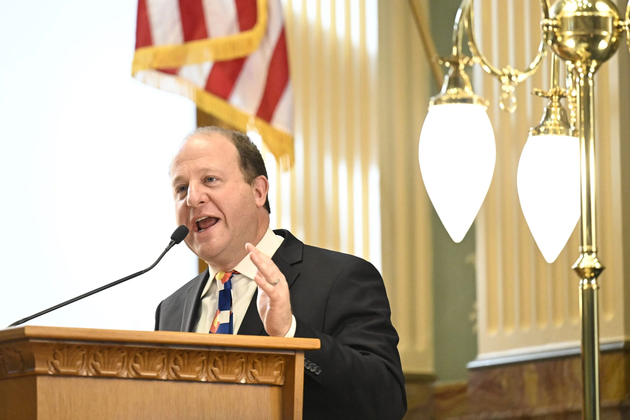 A man in a suit speaking at a podium with a microphone, with national flags and elegant lighting in the background. Eric Schaeffer of Fading West speaks before Congress about affordable housing crisis.