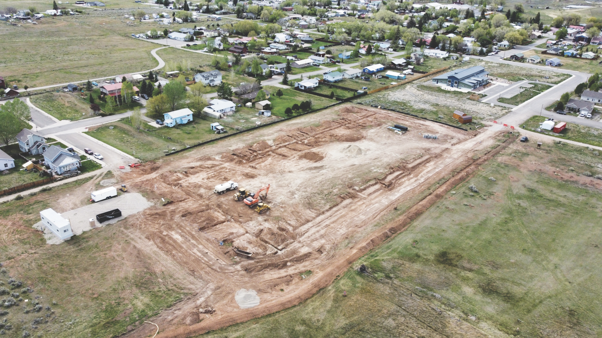 Aerial view of a Fading West modular housing multi-family construction site in a residential neighborhood with houses, trees, and roads, showing land being cleared and prepared for building.