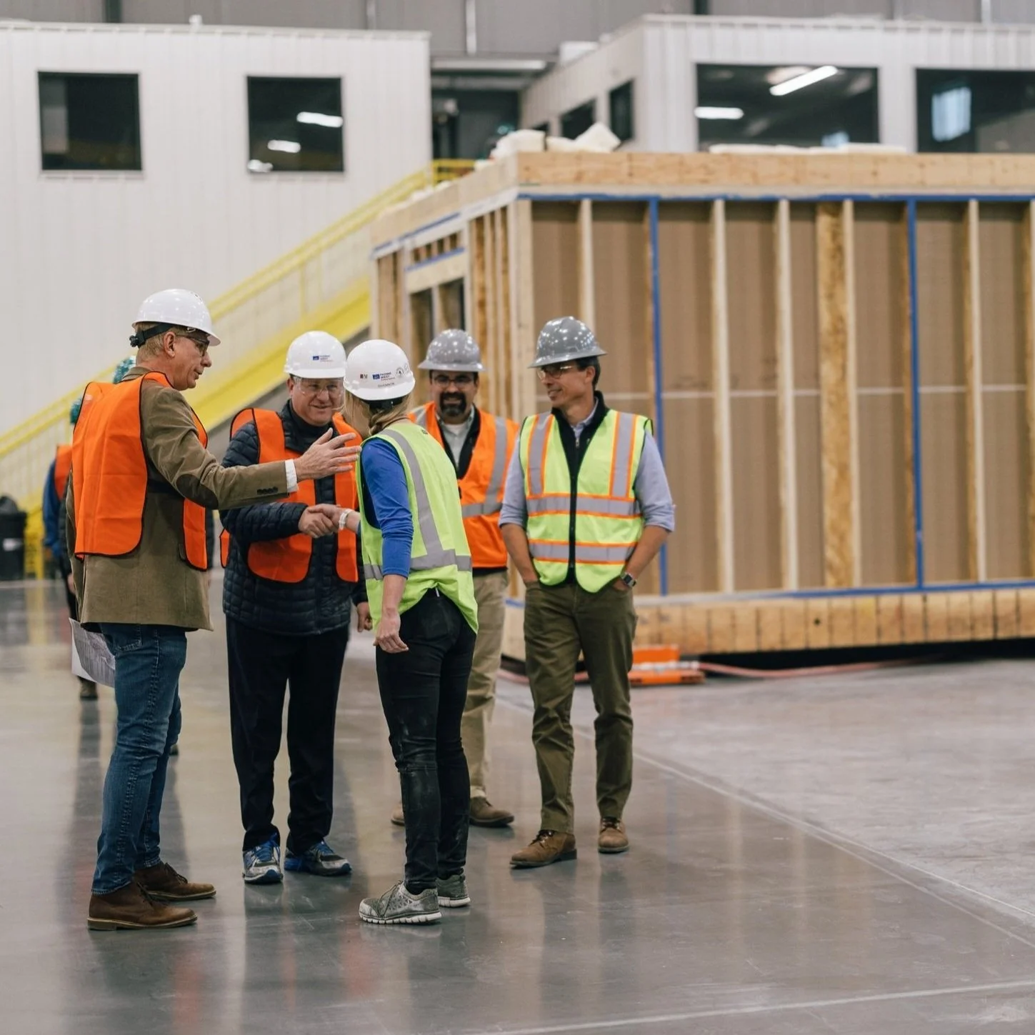 Group of construction workers and professionals wearing safety vests and hard hats in an indoor construction site, discussing near a partially built wooden structure at the Fading West modular housing factory in Buena Vista, CO