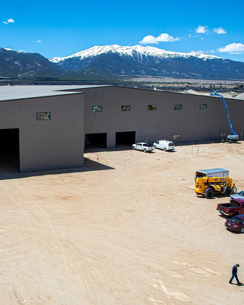 Fading West Construction site with a large gray warehouse, several parked vehicles, a person walking, and snow-capped mountains in the background under a blue sky.