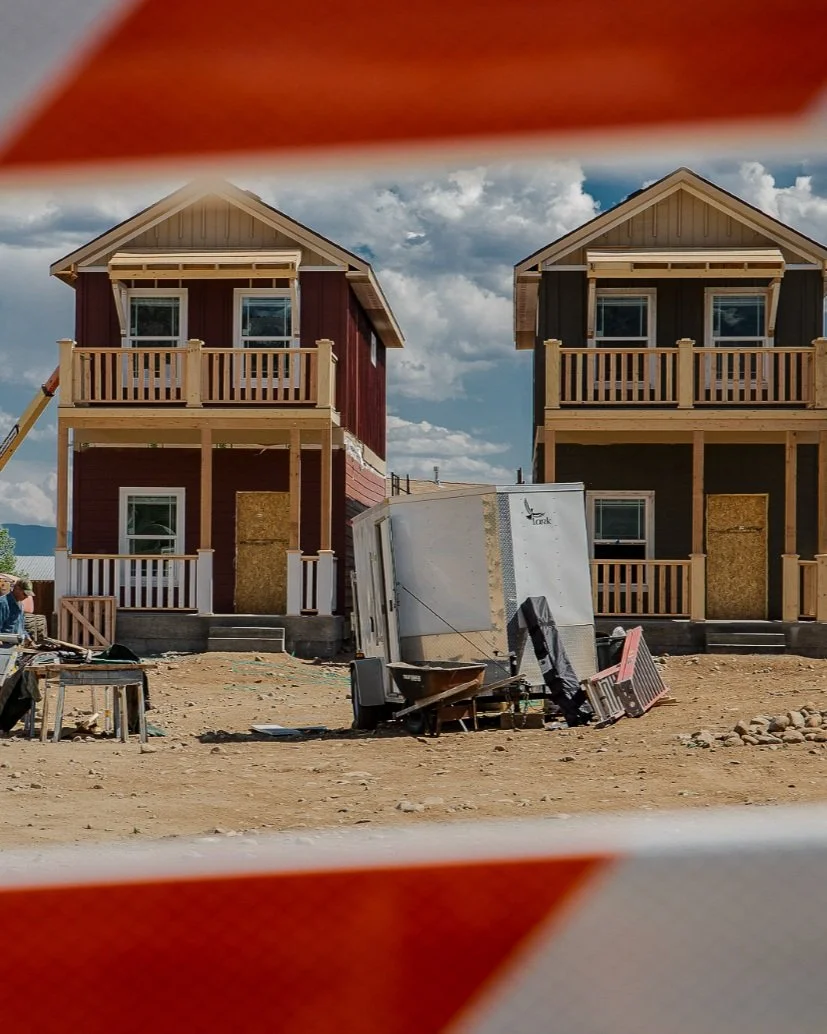 Two houses under construction with wooden balconies, surrounded by construction debris and equipment, under a partly cloudy sky.