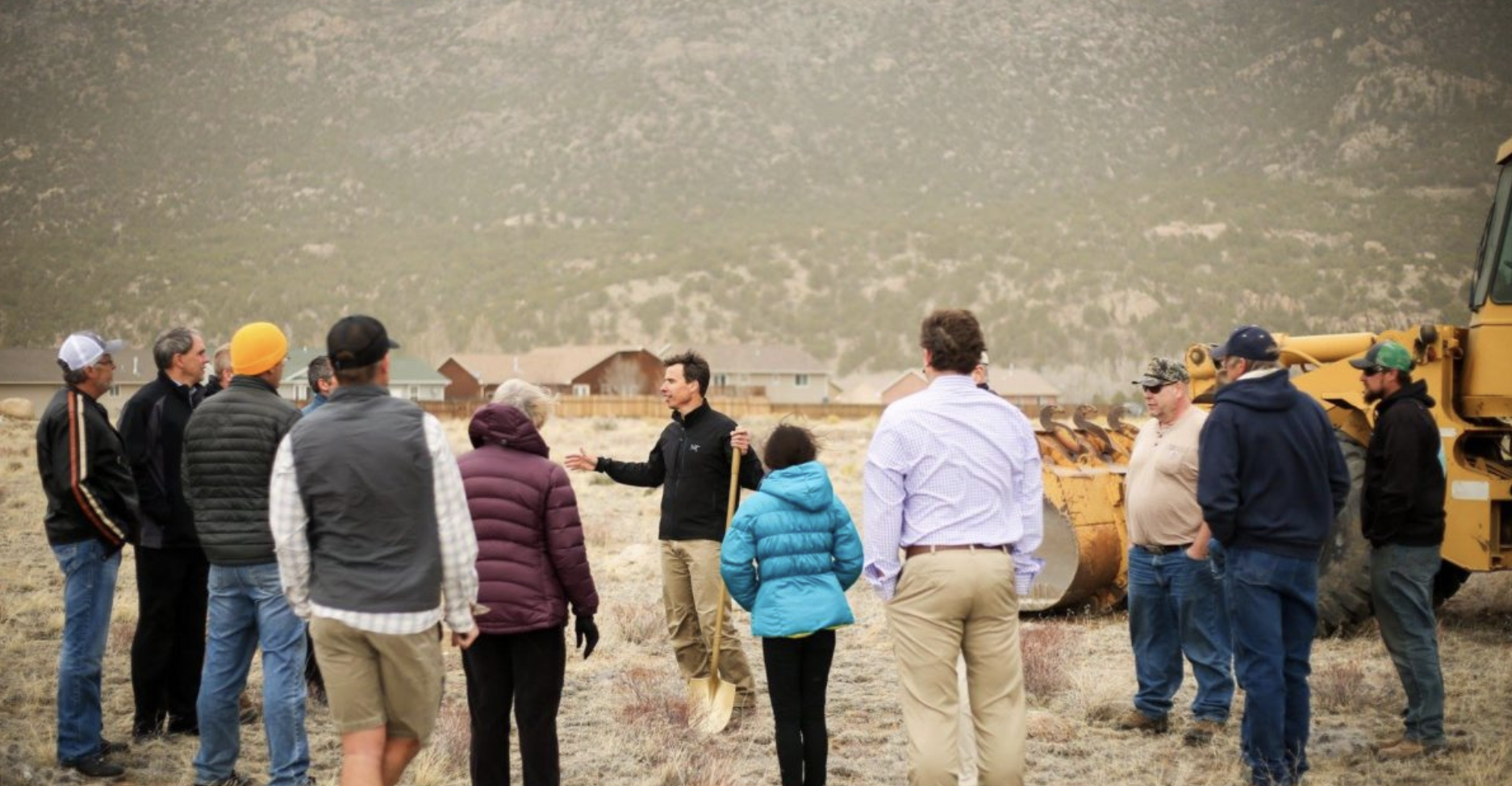 A group of people are gathered outdoors in a dry, grassy area with mountains in the background. They are listening to a man in black giving a presentation or speech. A yellow construction vehicle is partially visible on the right side of the image.
