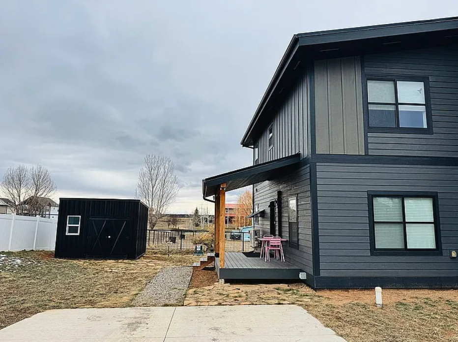 Two-story single-family modular blue house  by Fading West with a small porch, pink chairs, a black shed, and a backyard fence on a cloudy day.