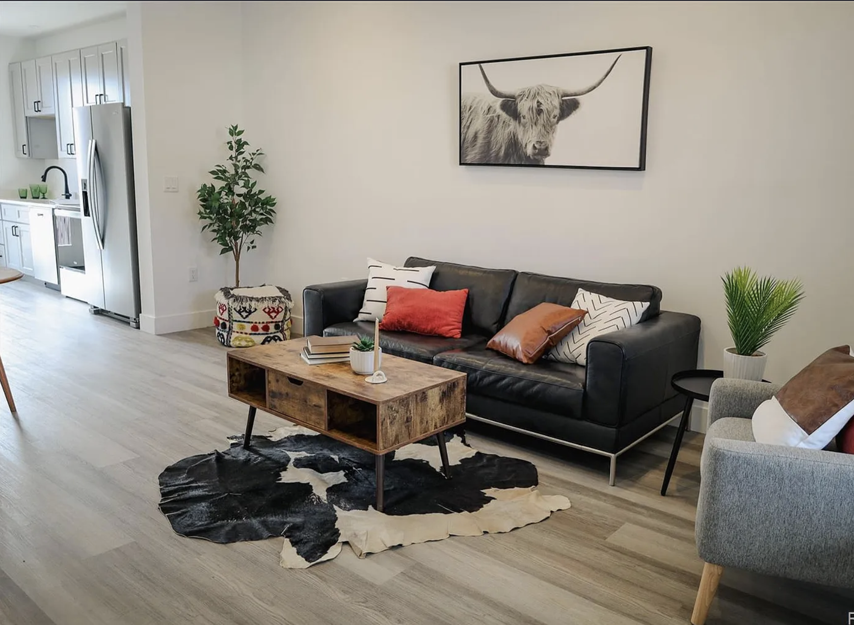 Interior photo of a Fading West single-family modular home featuring an open floorplan, Living room with black leather sofa, wooden coffee table, gray armchair, indoor plants, framed highland cow picture on the wall, and decorative pillows,