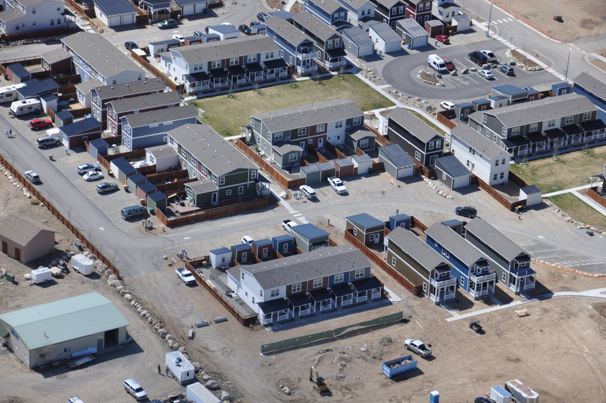 Aerial view of a modular home residential neighborhood with multiple modern modular home townhouses, parking lots, and some construction activity. The neighborhood is organized with small yards and fenced areas.