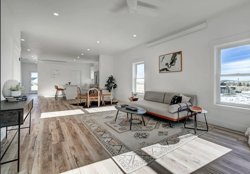 Interior photo of a Fading West Modular Single Family Home featuring a bright, modern living room and dining area with white walls, hardwood floors, a beige sofa, a patterned area rug, a black coffee table, and a dining table with chairs.