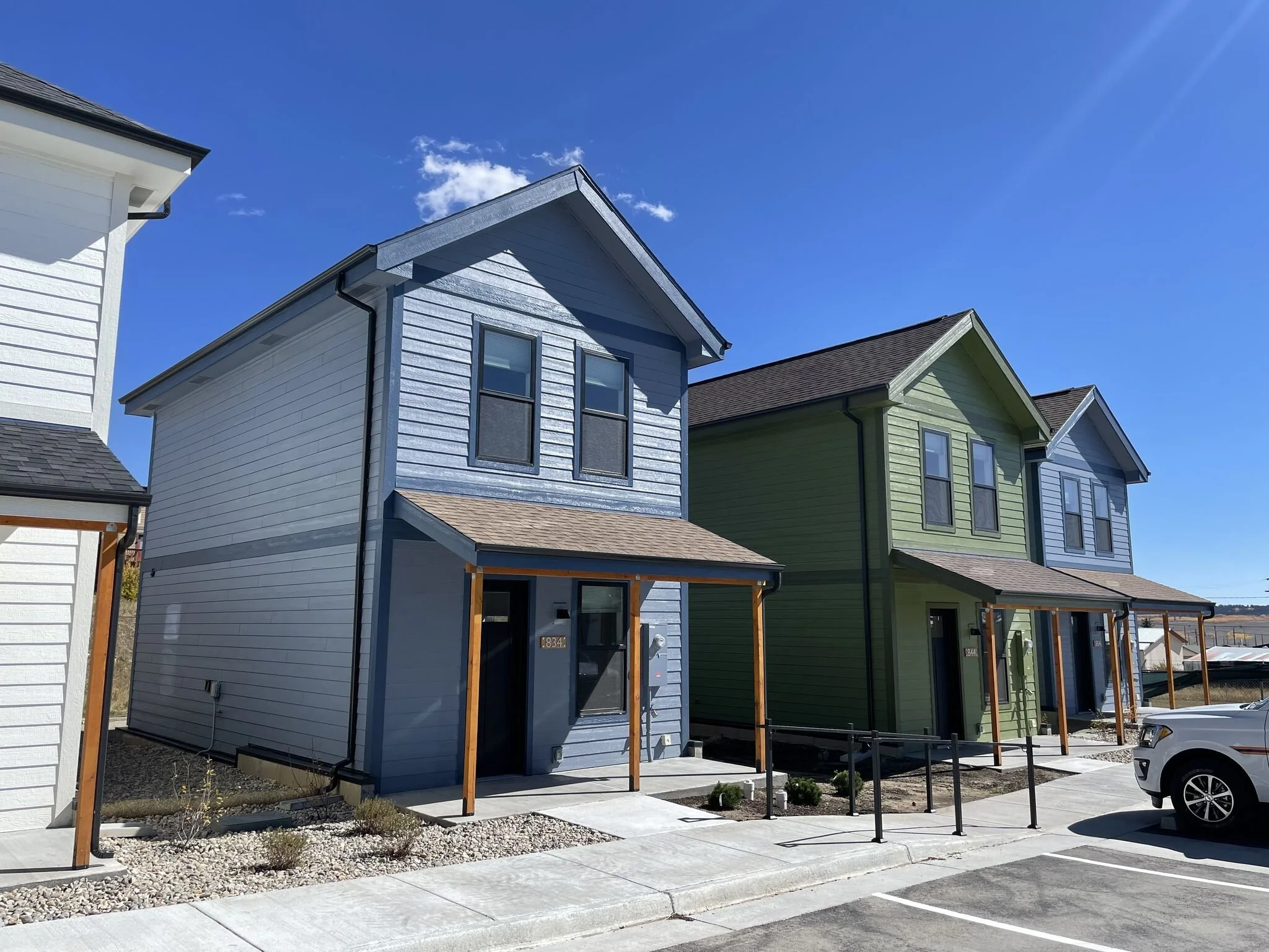 A row of newly built single family modular houses with colorful exteriors, including blue, green, and light gray, under a bright blue sky with a few clouds. The houses have modern designs with large windows