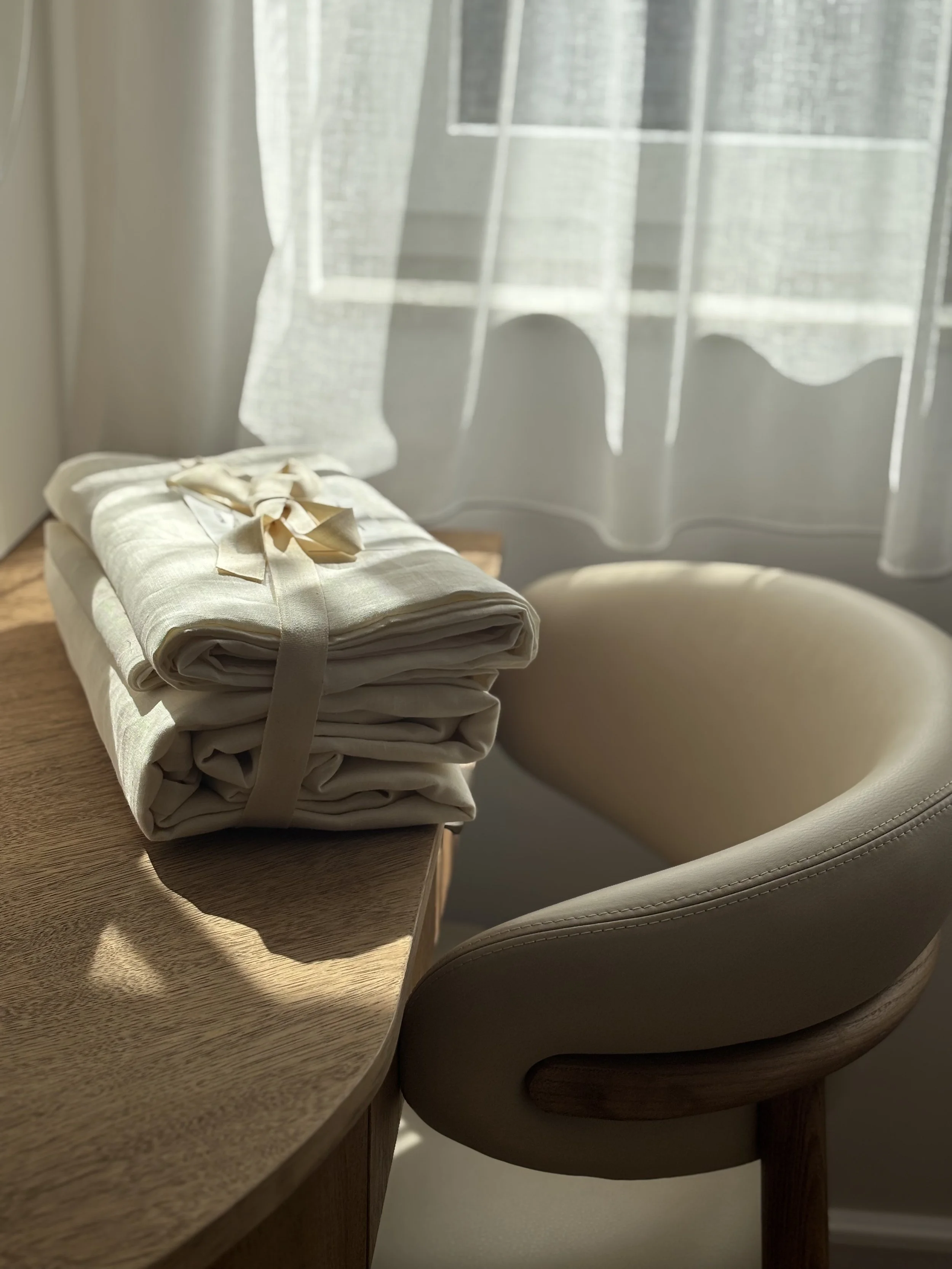 A neatly folded cream-colored fabric with a ribbon tied on top, placed on a wooden table next to a curved beige chair with wood legs, in front of a window with sheer white curtains and sunlight casting shadows.