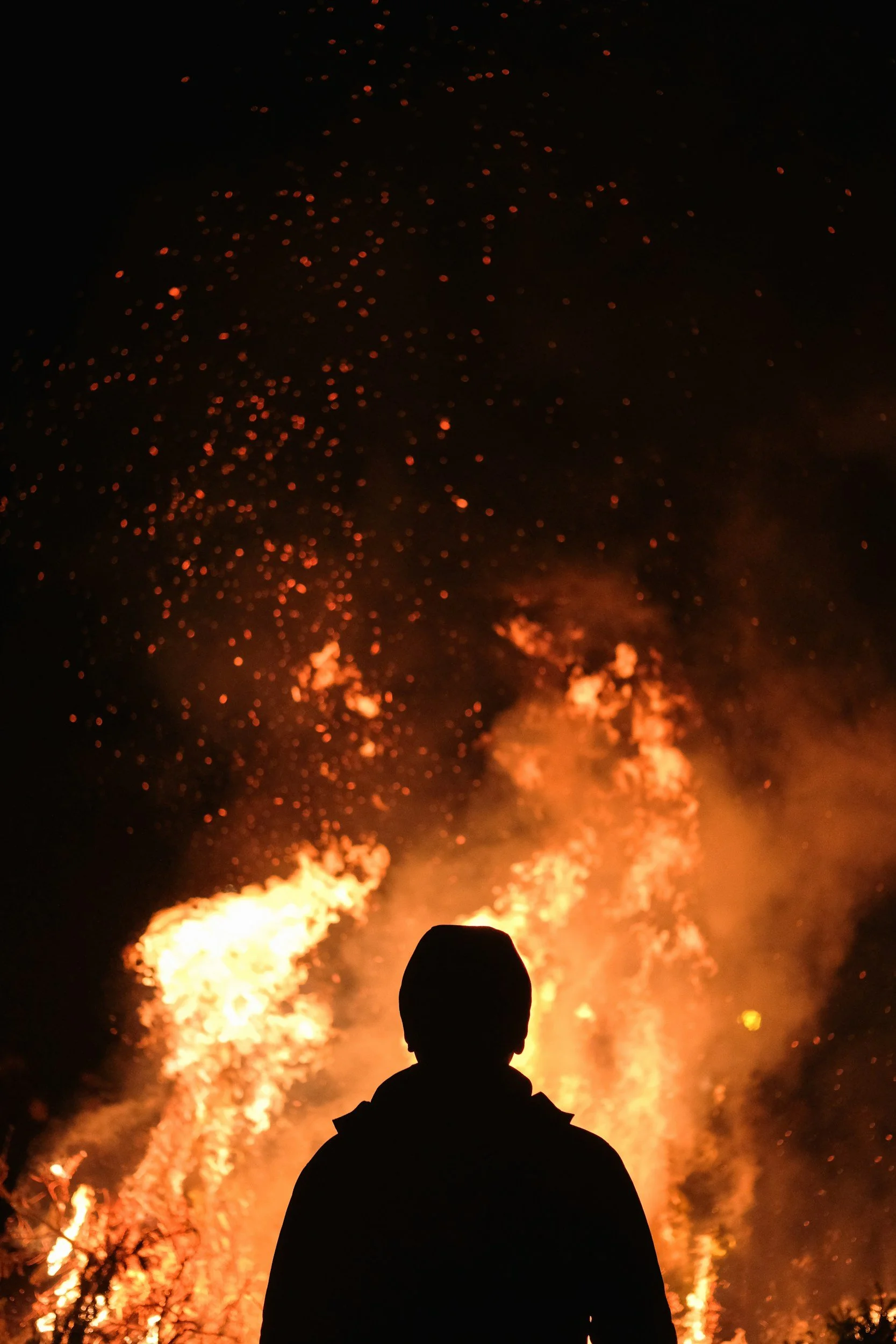 Person standing and observing a large fire with flames and sparks at night.