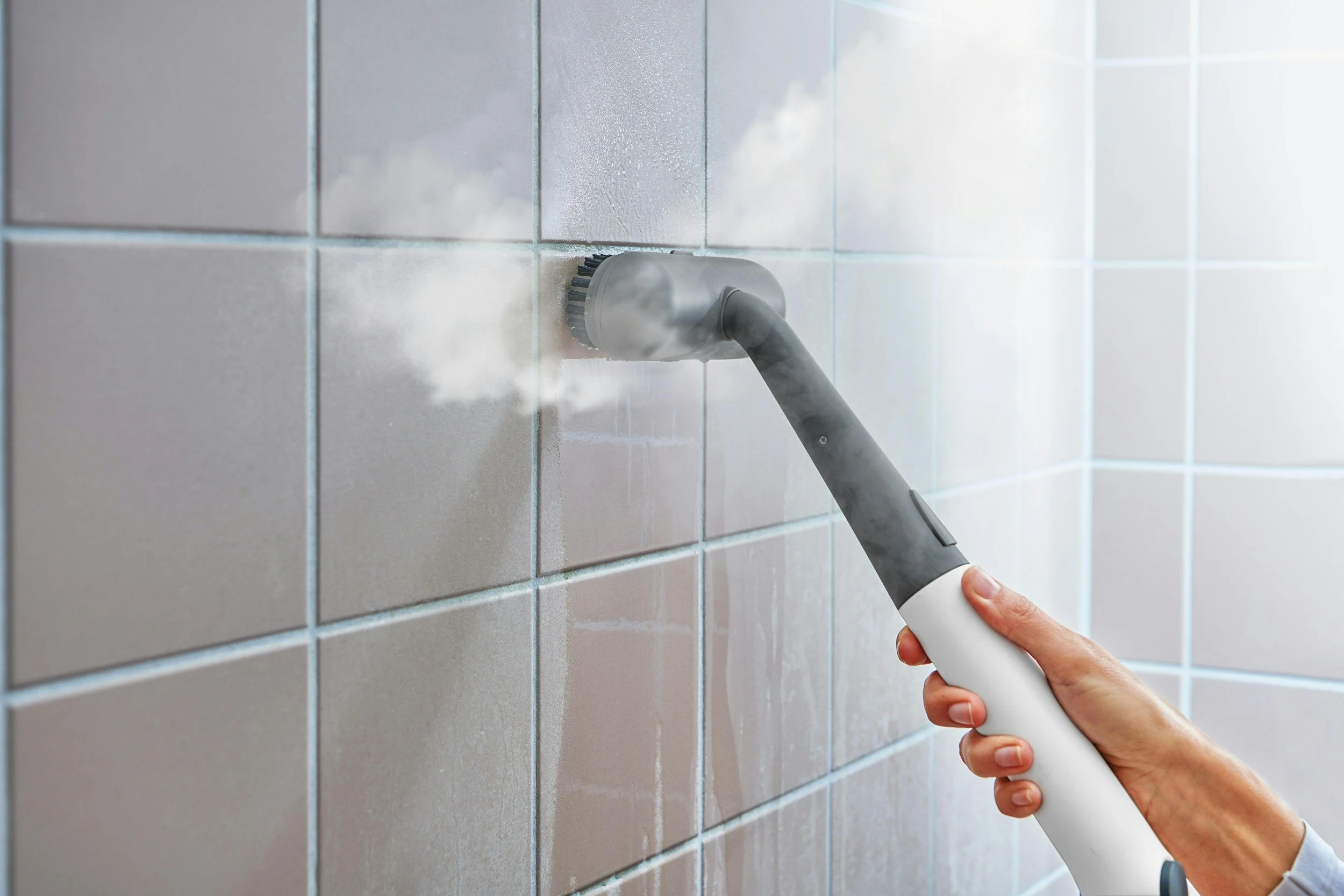 A person cleaning light gray tiled wall with a handheld steam cleaner.
