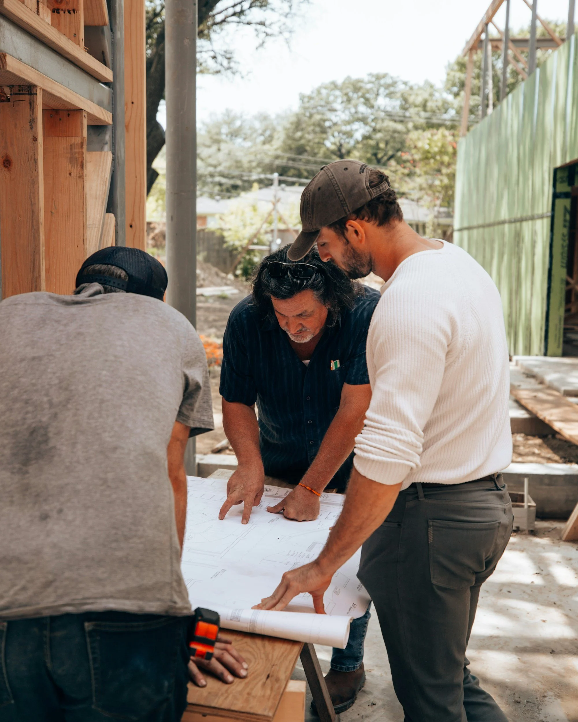 Three men reviewing blueprints at a construction site, with wooden framing and green construction fencing in the background.