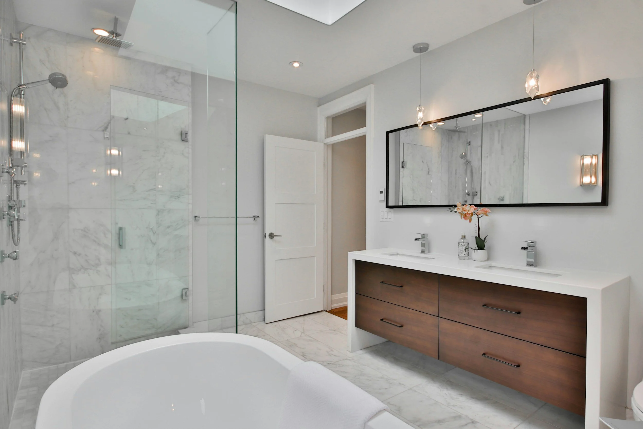 Modern bathroom with a glass-enclosed shower on the left, white marble walls and flooring, a white vanity with a dark wood cabinet, and a large rectangular mirror above the sink. Decorative pendant lights hang from the ceiling, and a small potted plant with pink flowers is on the vanity.