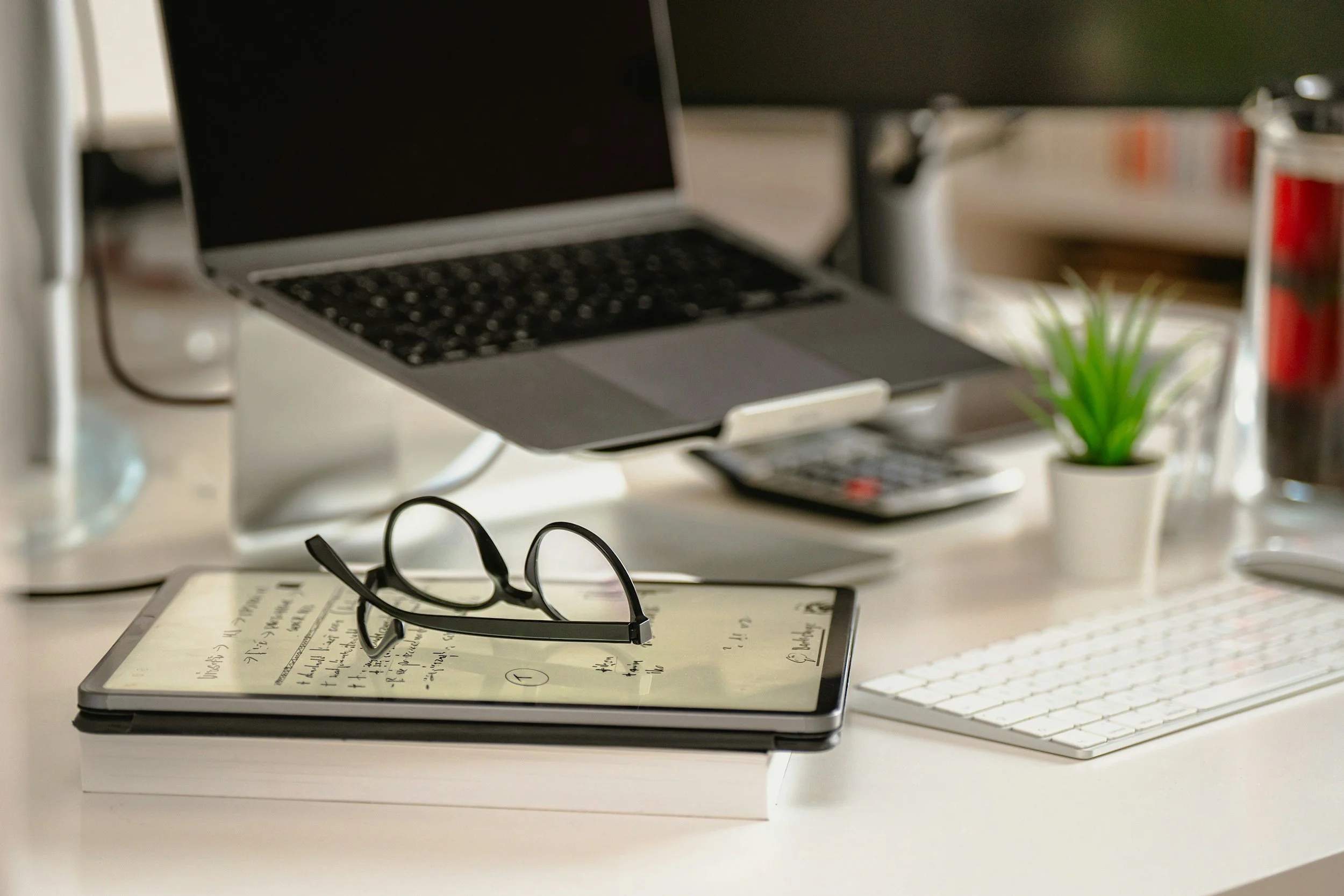 A laptop on a desk with glasses resting on an electronic tablet displaying handwritten notes, with a pair of glasses placed on the tablet. A small potted plant and various office supplies, including a keyboard and a remote, are visible in the background.