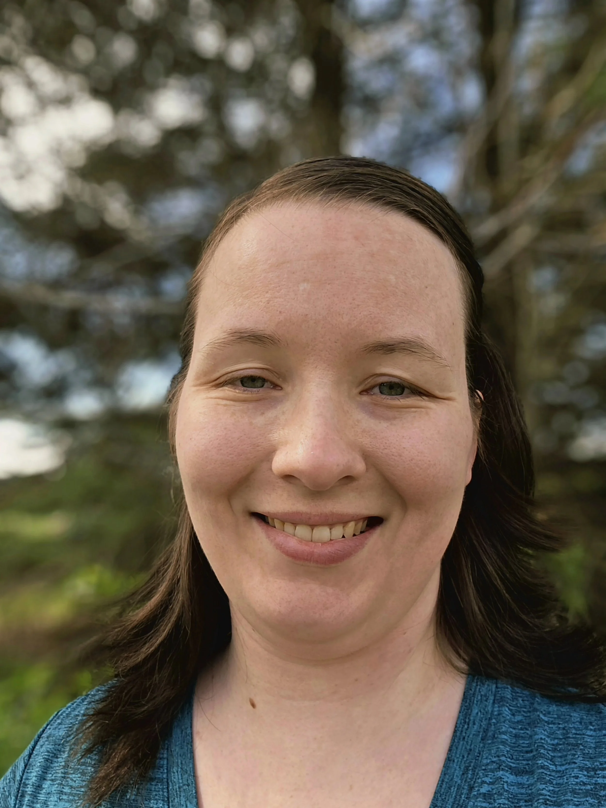 Close-up of a smiling woman with dark hair and light skin outdoors, with trees and blurred background.