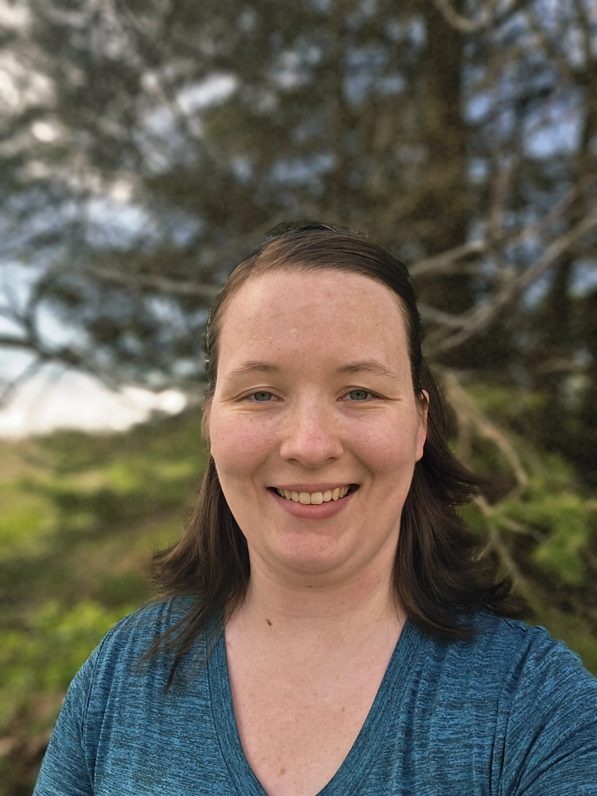 Close-up of a woman smiling outdoors with trees and sky in the background.