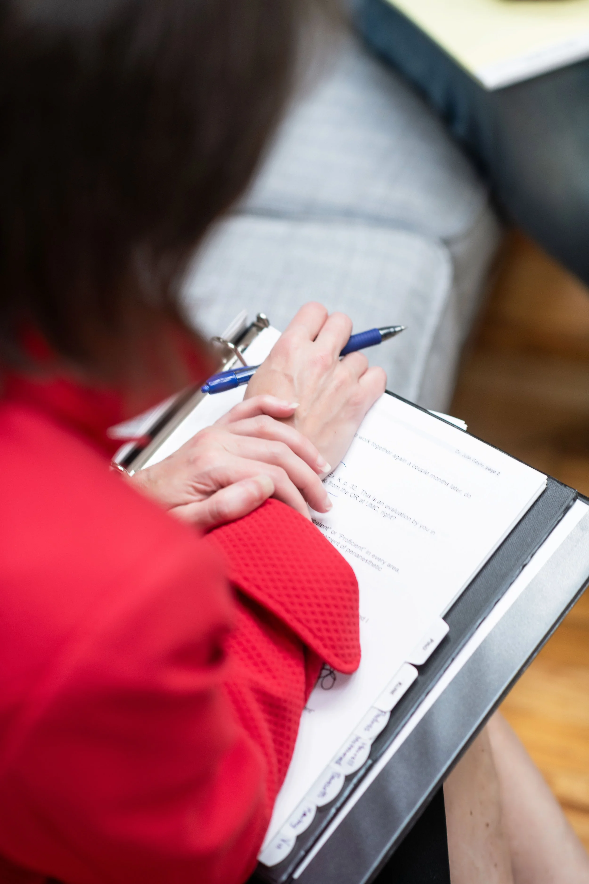 A person wearing a red garment is sitting at a table, holding a blue pen and writing on a clipboard with papers. The background shows a gray cushioned chair and a window.