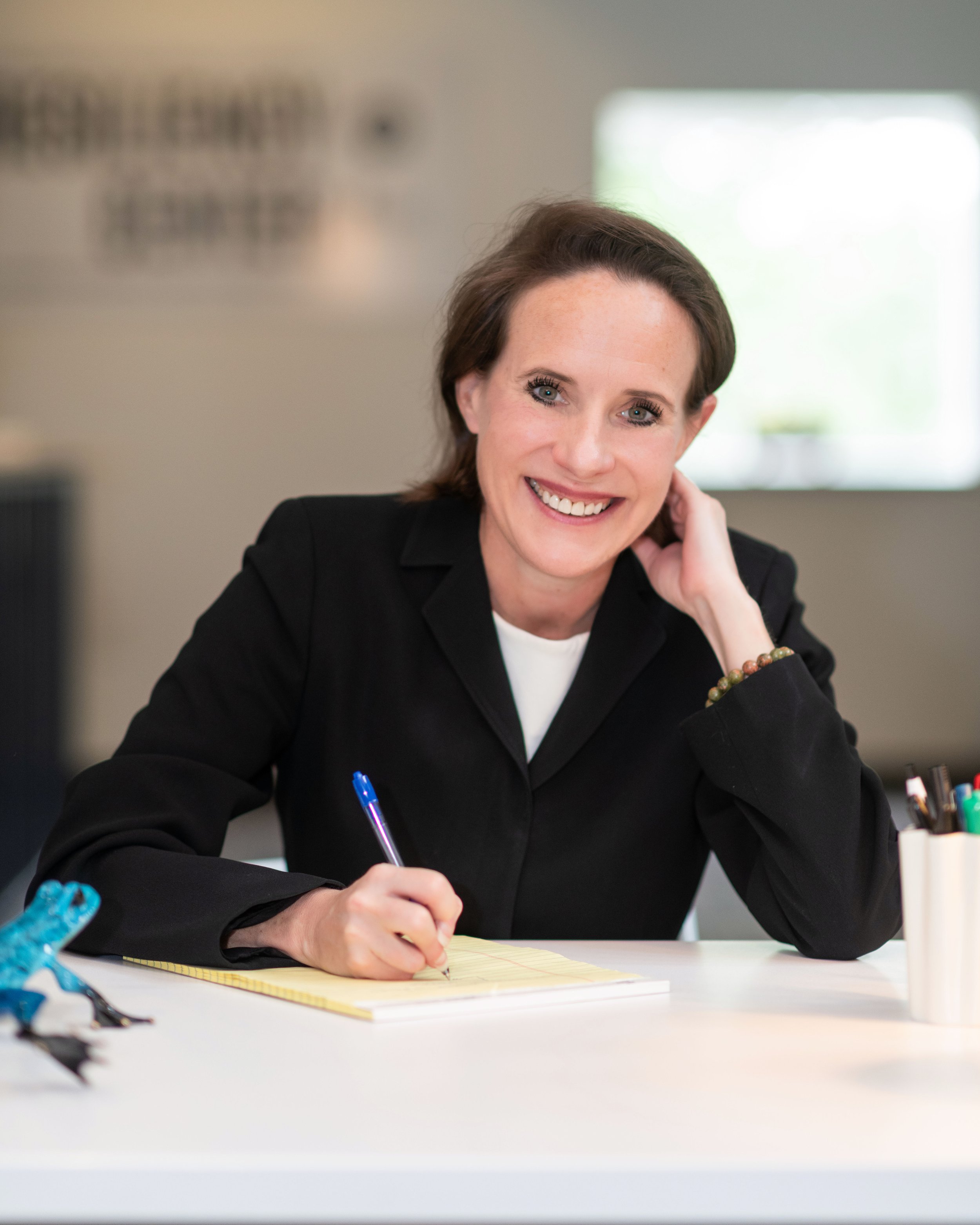 A woman with short brown hair and blue eyes smiling while sitting at a desk, writing on a yellow notepad with a blue pen. She is wearing a black blazer and has a beaded bracelet on her right wrist. The background is blurred with a window visible.