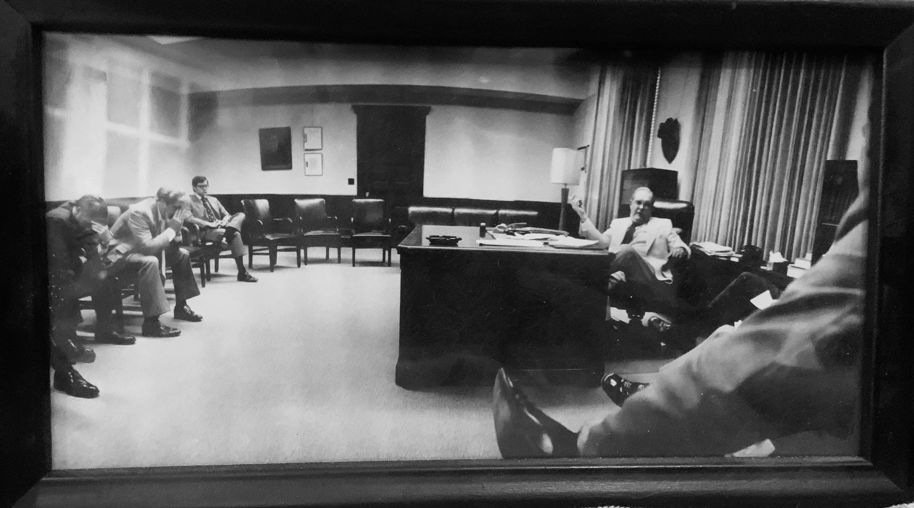 Black and white photograph of a meeting room with five men seated, four on the left and one at a desk on the right. The man at the desk is holding a telephone receiver, surrounded by documents and office supplies, suggesting a formal or business setting.