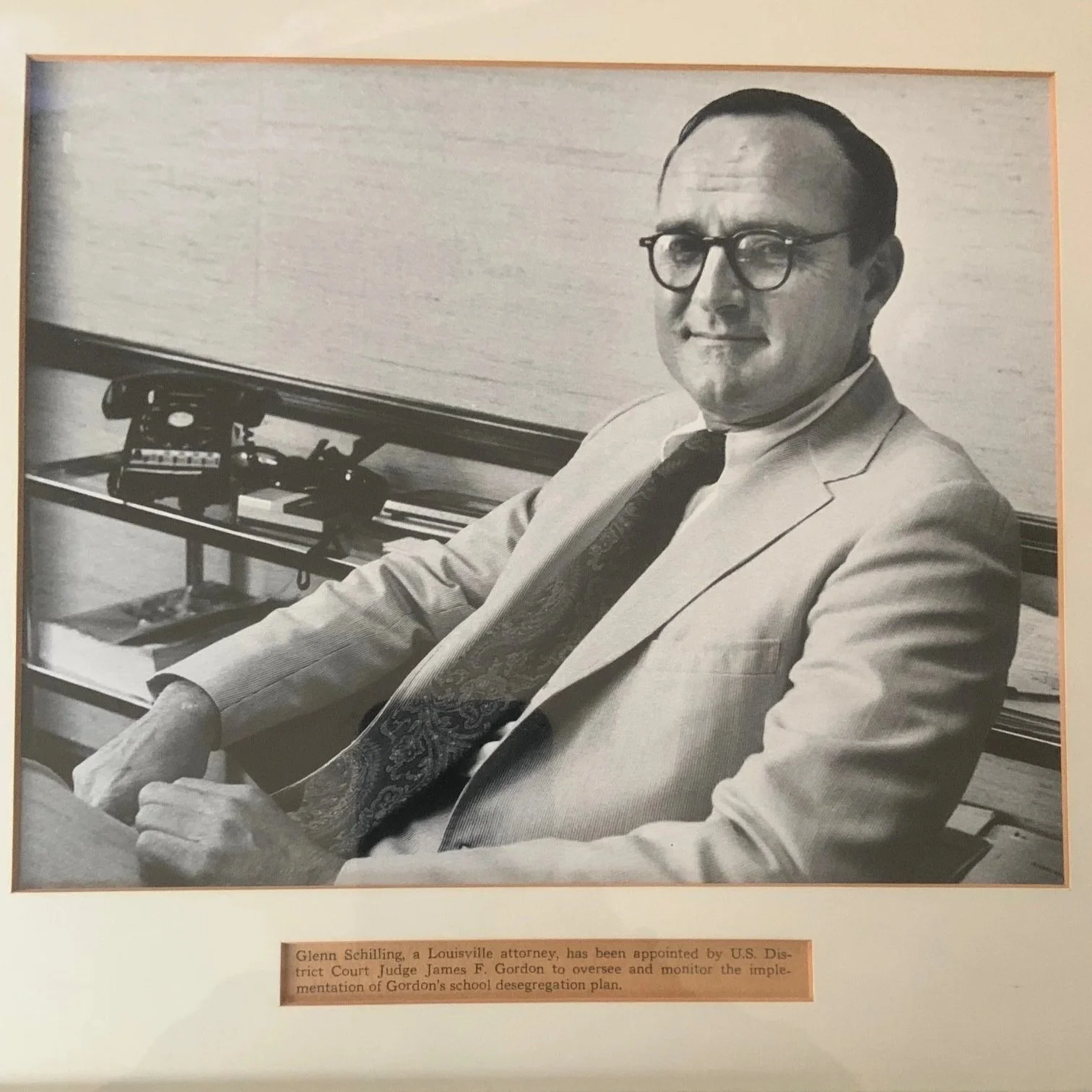 A black and white photograph of Glenn Schilling, a Louisville attorney, sitting at a desk. He is wearing glasses, a light-colored suit, a white shirt, and a patterned tie. There is a telephone and some books or papers on a shelf behind him. A caption below the photograph states, "Glenn Schilling, a Louisville attorney, has been appointed by U.S. District Court Judge James F. Gordon to oversee and monitor the implementation of Gordon’s school desegregation plan."