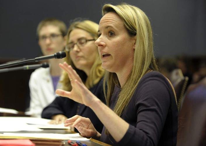 A woman is speaking into a microphone during a formal meeting or hearing, with others listening beside her.