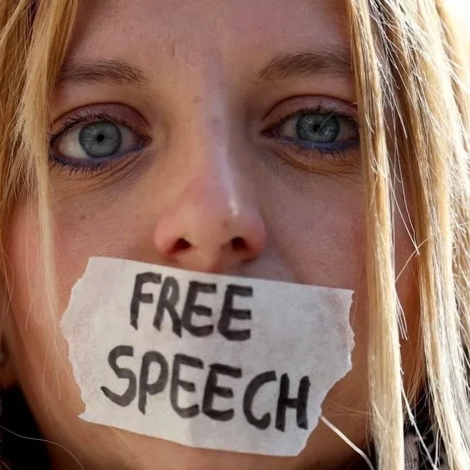 A woman with blue eyes and blond hair holding a torn piece of paper with 'FREE SPEECH' written on it over her mouth.