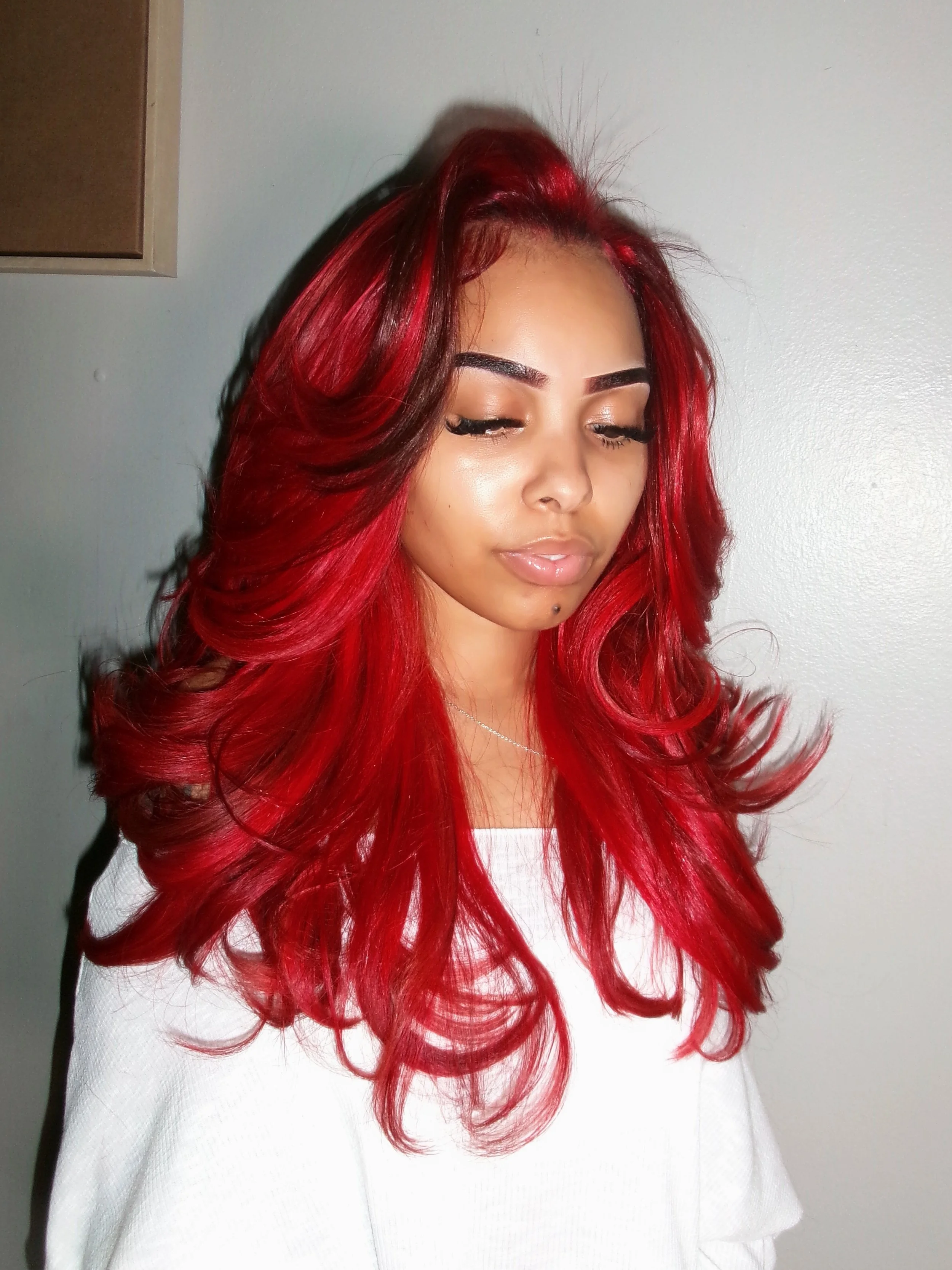 A woman with long, wavy red hair and closed eyes, wearing a white top, standing against a plain light-colored wall.