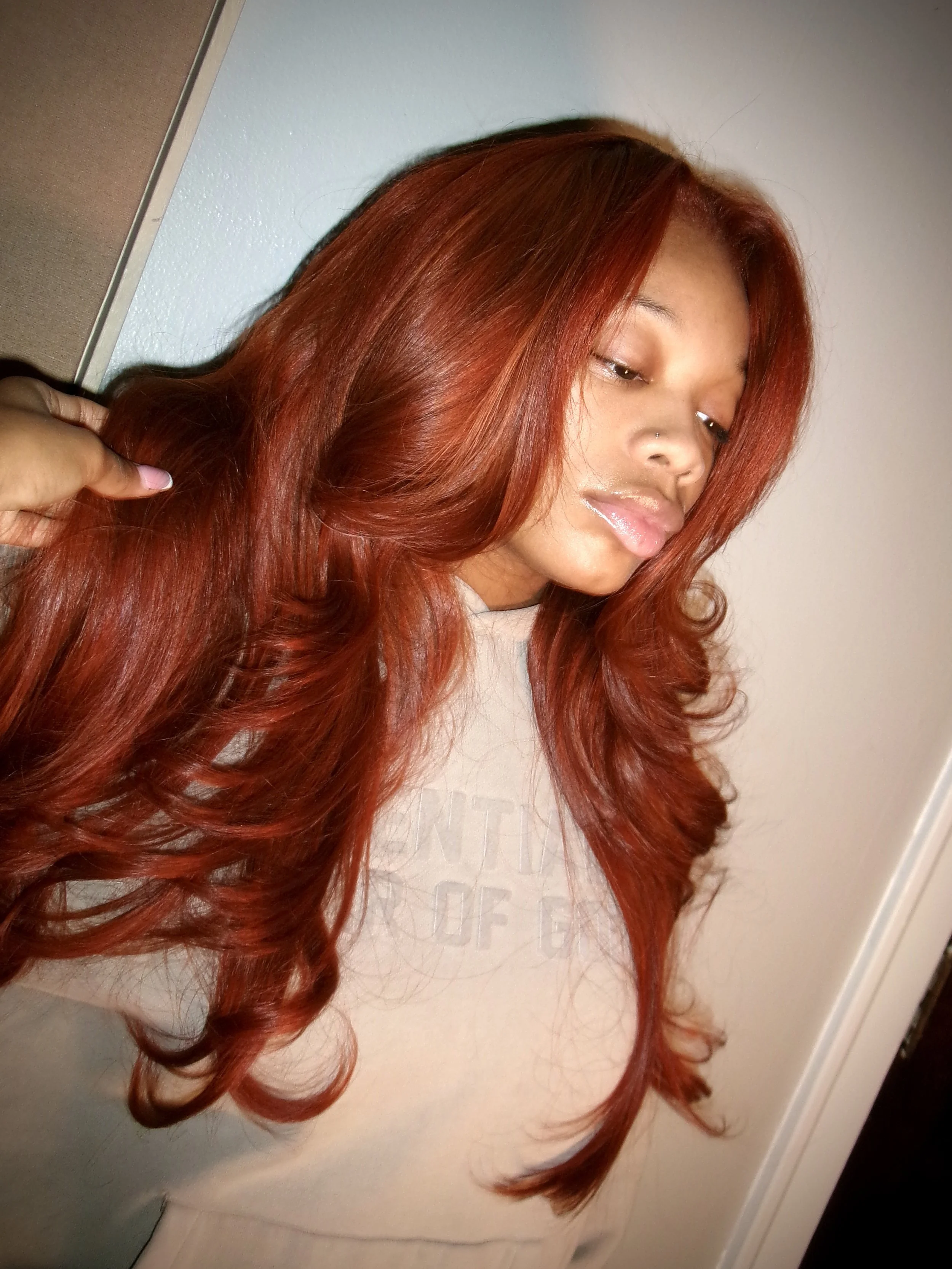 A young woman with long, wavy, red hair taking a selfie in a mirror, standing against a plain wall and dresser.